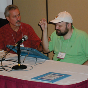 Picture of Tracy presenting with another man. They are sitting behind a table and there is a microphone on the table.