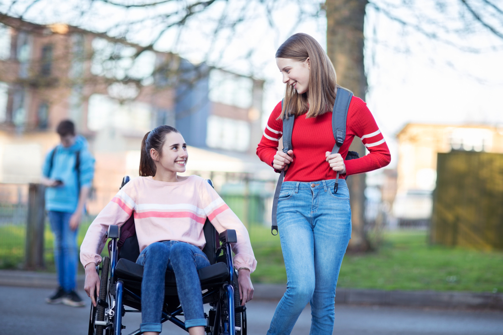 Two girls looking at each other and smiling. One is in a wheelchair. The other is standing and wearing a backpack.