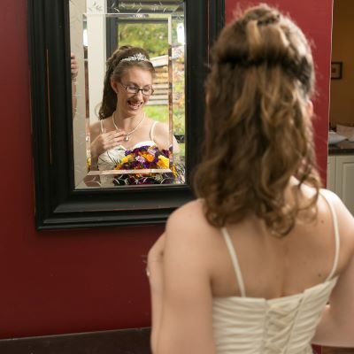 A woman in a wedding dress touching her necklace and looking down, smiling. The photo is from behind but the woman's face is visible in a mirror.