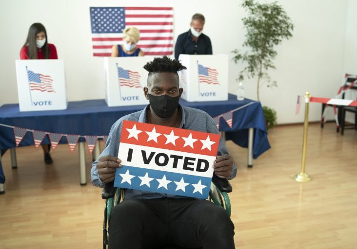 Young man in a wheel chair holding an American flag at election center, wearing a face mask